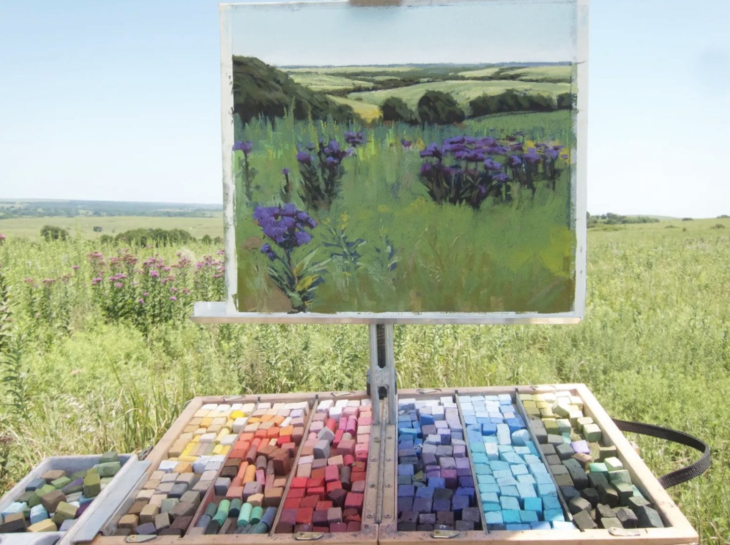 Nature sketching example with pastels. You can see a pastel drawing of a springtime landscape and an easel with a buch of colors of pastel all layed out.